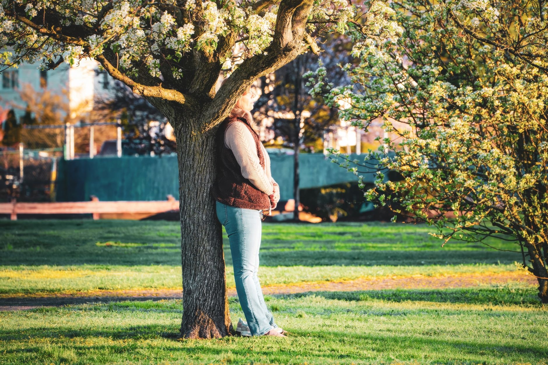 A woman leans against a flowering tree in a sunny park, enjoying a peaceful spring day outdoors.