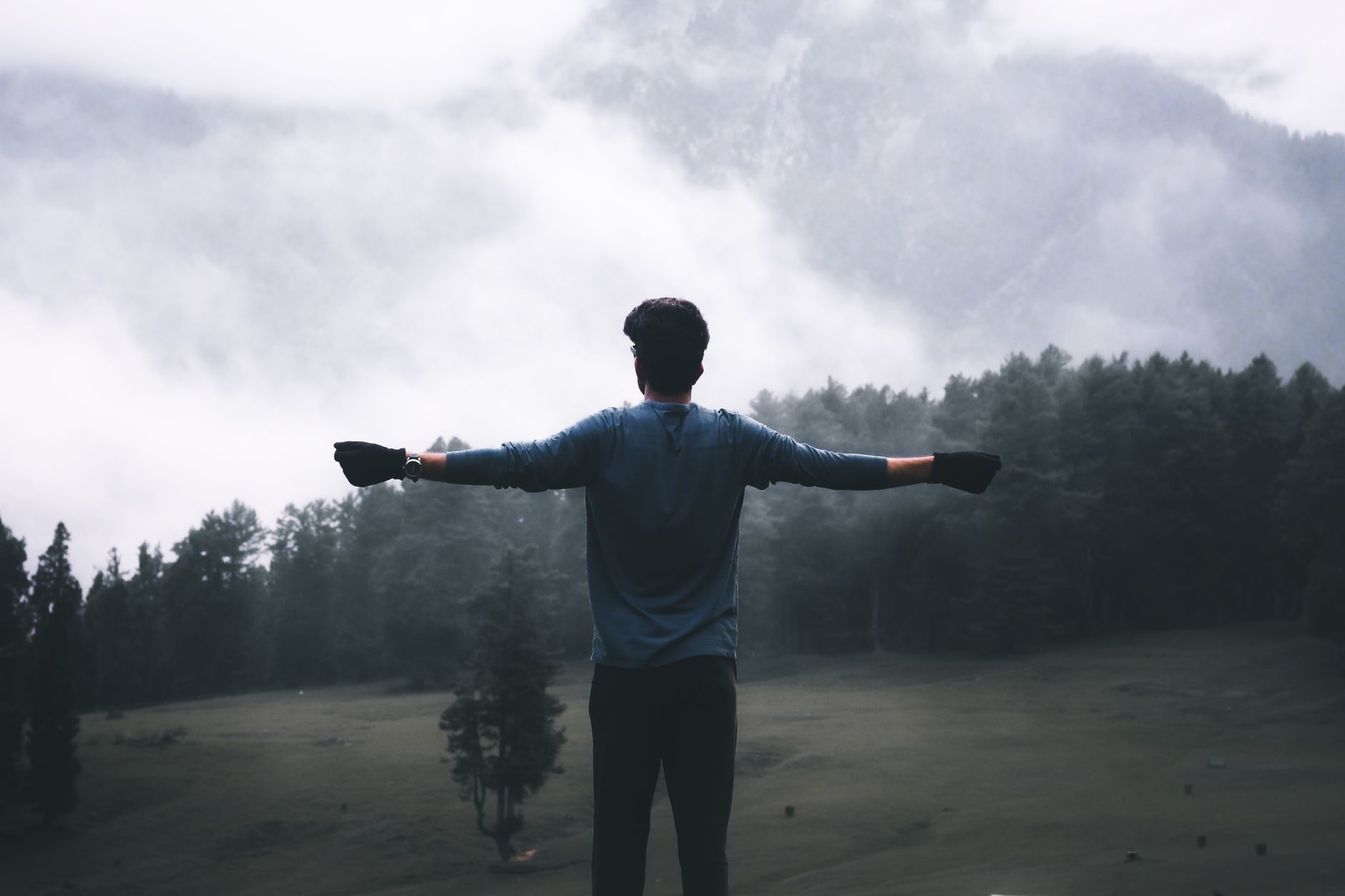 A man stands with outstretched arms, taking in the foggy forest and mountainous view.