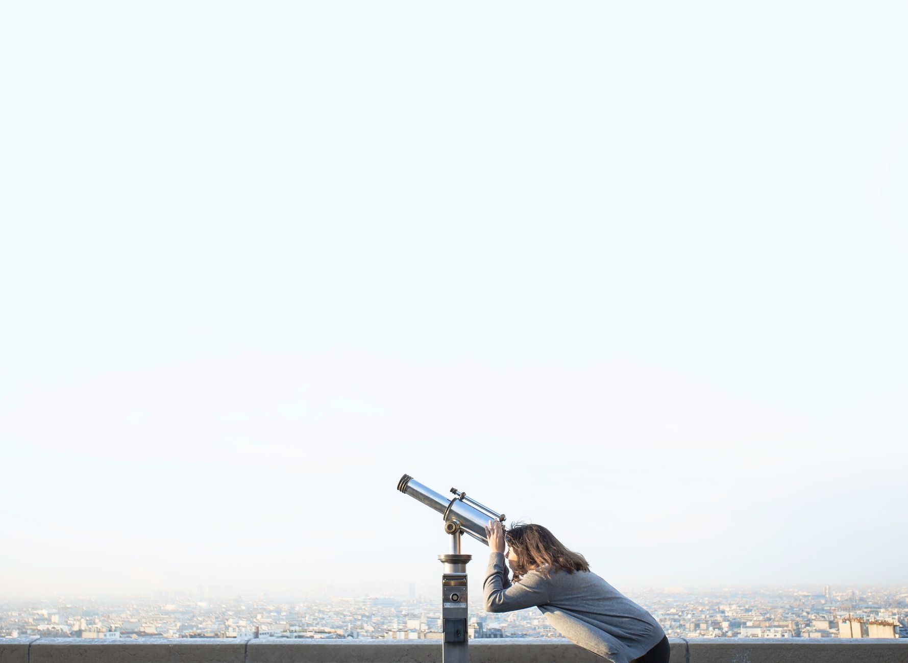 A woman peers through a coin-operated telescope overlooking a city skyline.
