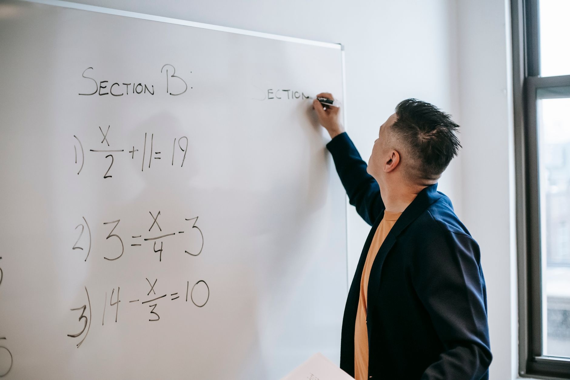 Side view of focused teacher in jacket writing equations on whiteboard in light room in daylight