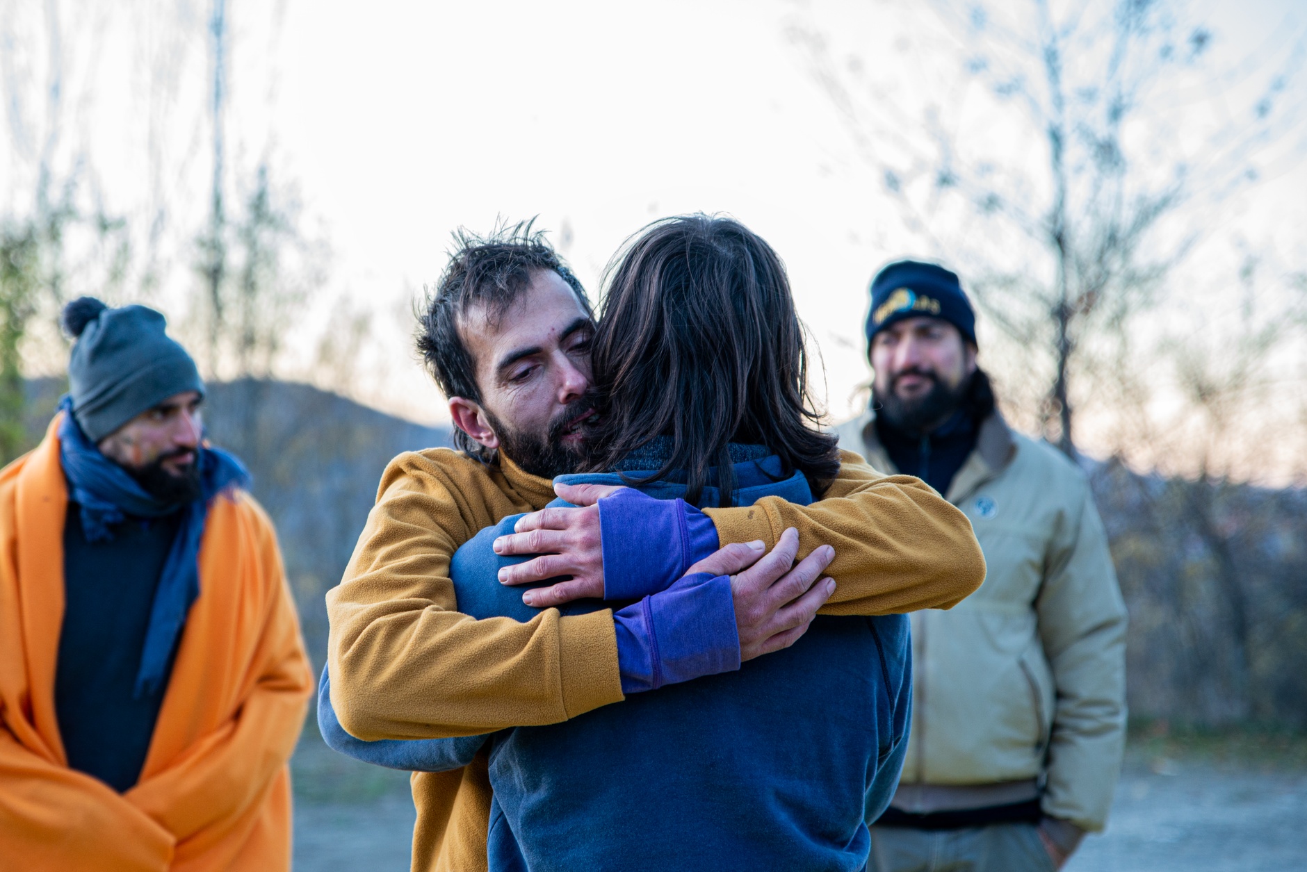 A heartfelt hug between friends outdoors during a cool, bright day.